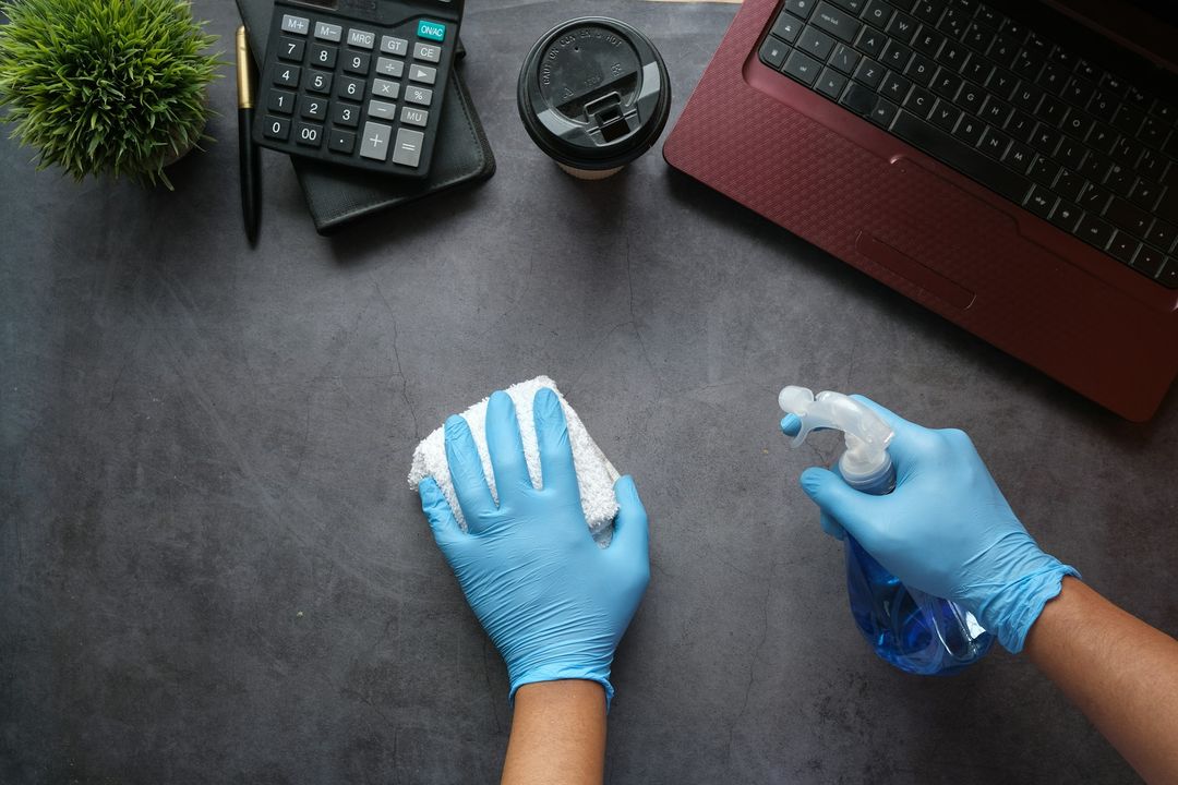 Schreibtisch wird mit Handschuhen und Reinigungsspray abgewischt